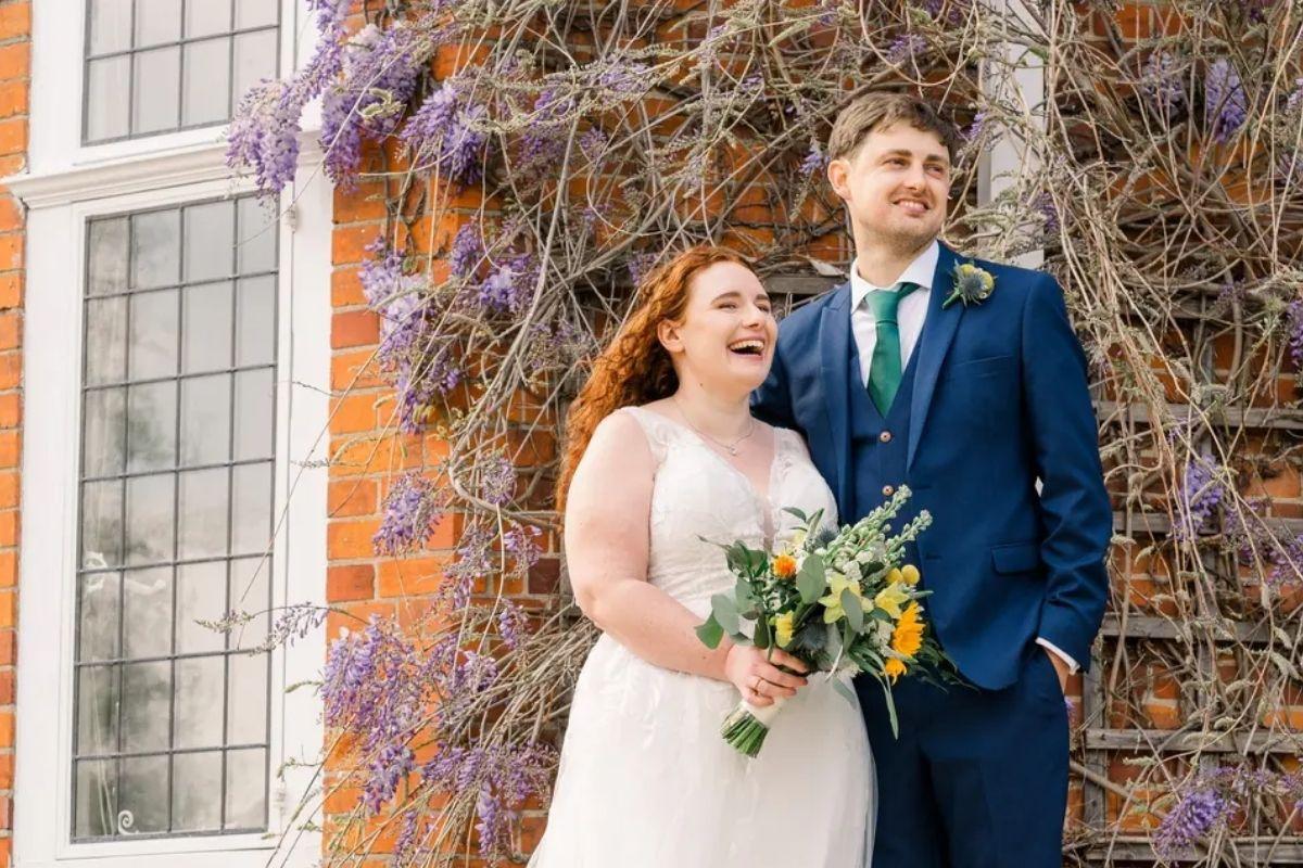 a sweet bride and groom pose on their wedding day outside their venue with lavendar coloured vines and plants behind them