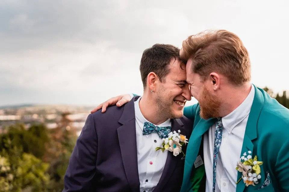 Two grooms embrace and smile at each other on a hill overlooking trees and a city