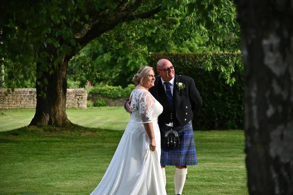 Bride and kilt-wearing groom pose for a picture outside by trees