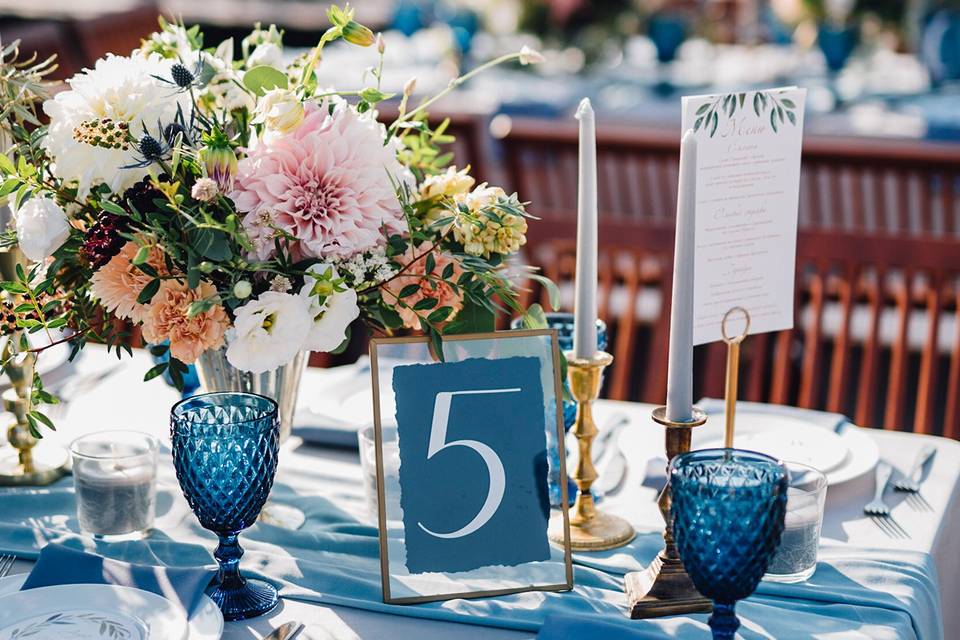 Wedding table setup with a lush centrepiece of flowers, and a table number '5' sign on acrylic in a gold frame