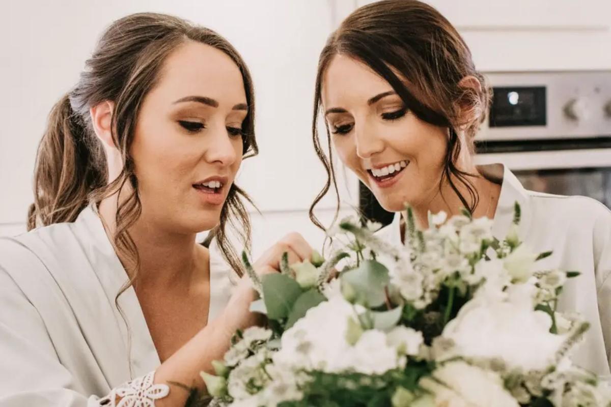 a bride and her bridesmaid wearing bridal party robes and looking at their bouquets with wavy ponytail hair - aura