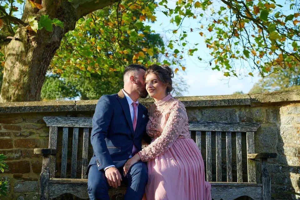 Bride and groom embrace on a bench in front of a stone wall and tree