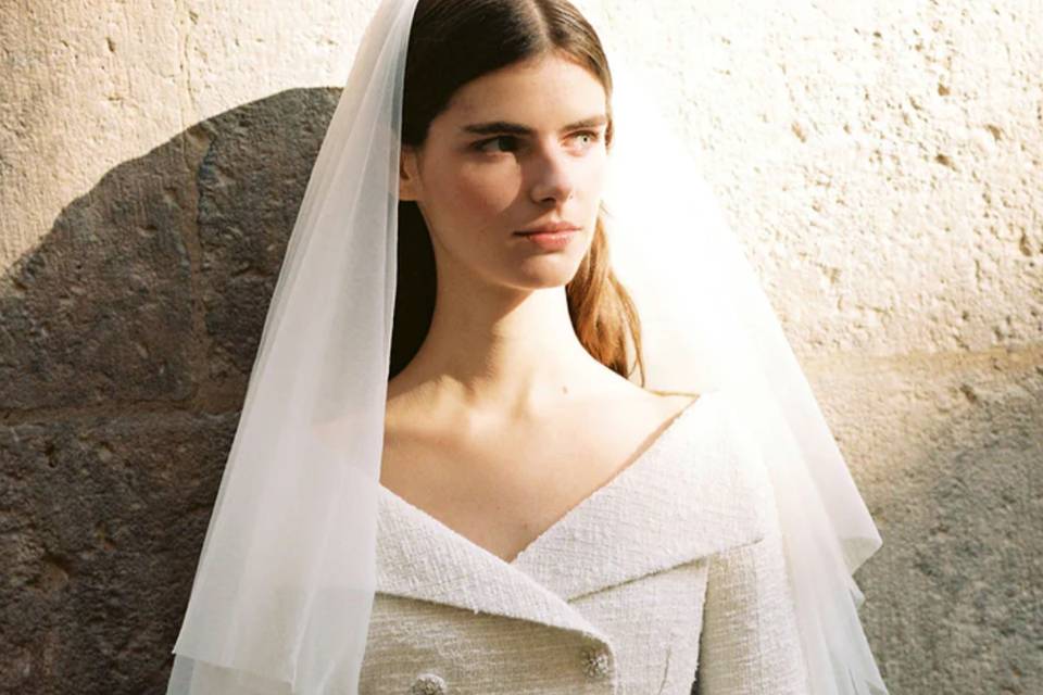 a bride stands against a wall in a registry office wedding dress blazer and veil looking to the right