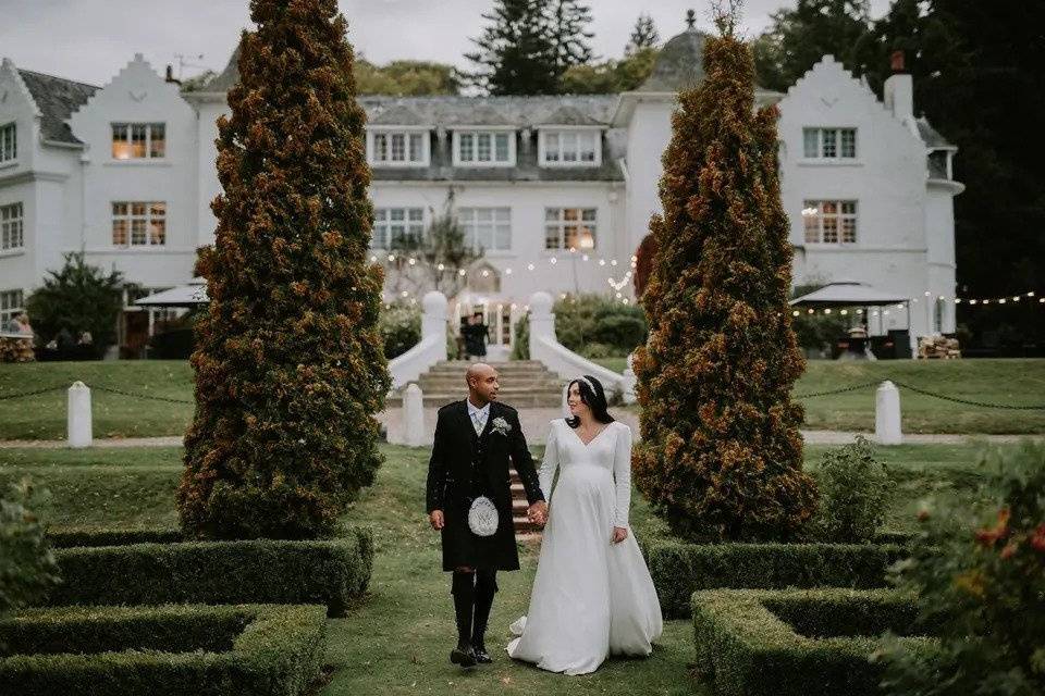 A bride and groom wearing a kilt walk through the manicured gardens in front of a white stately home