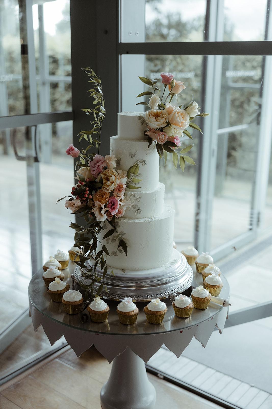 A white wedding cake dressed in flowers and surrounded by white cupcakes