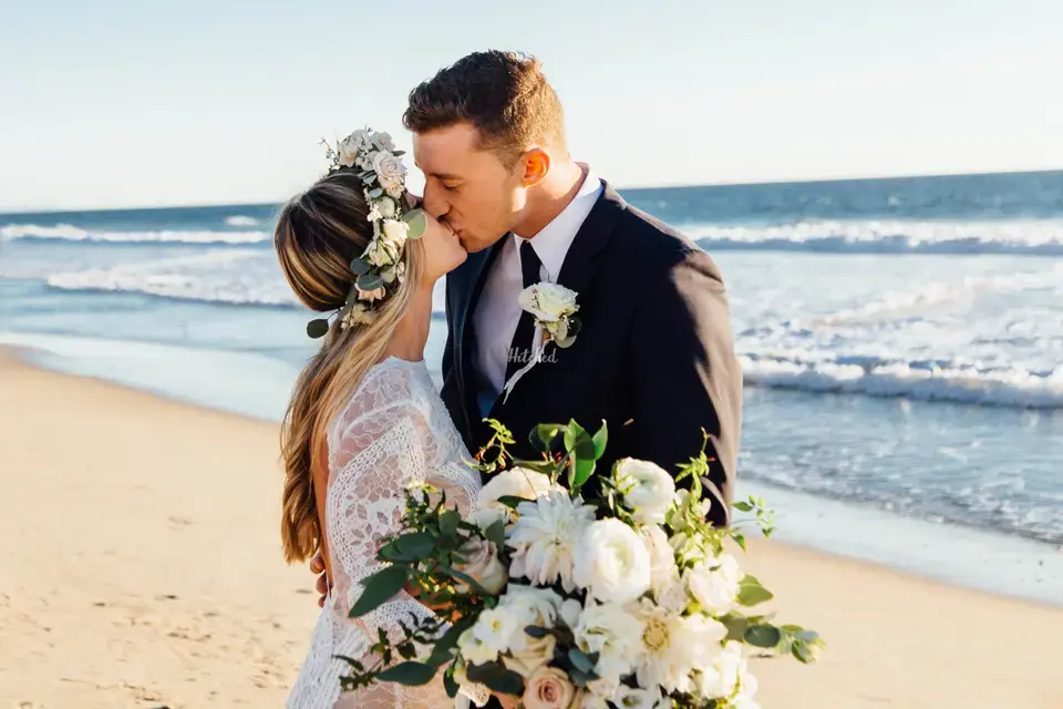 Boho style couple with a bride wearing a flower crown kissing on a beach next to the shore