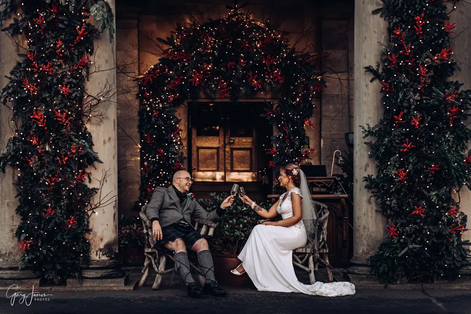 A groom in a kilt and traditional scottish formalwear sitting down outside a wedding venue with his bride, toasting with champagne. The venue is decorated with greenery and red bows and fairy lights for Christmas