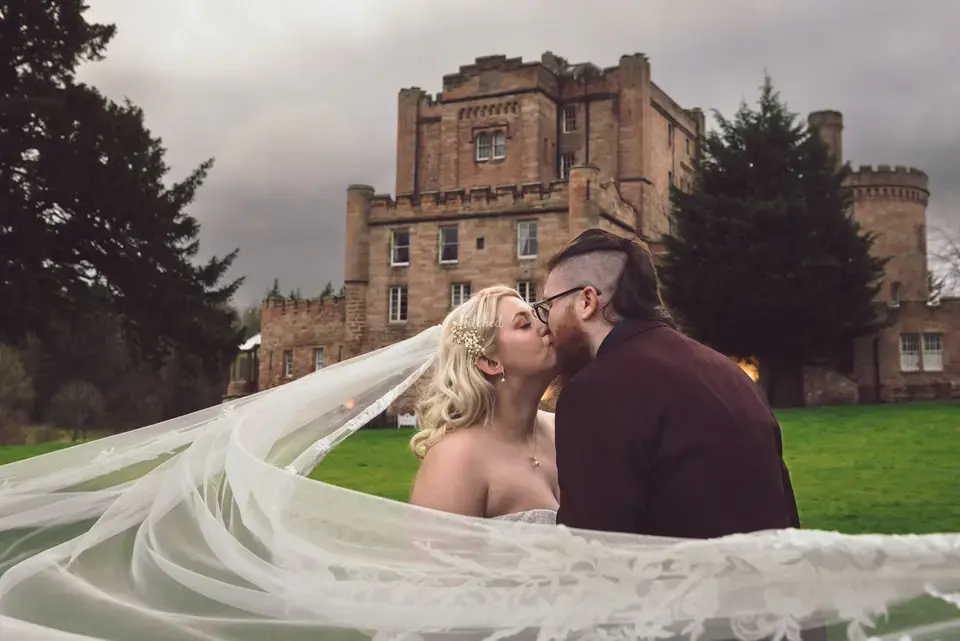 Couple kissing outside of a scottish wedding venue as the bride's veil is swept around them by the wind