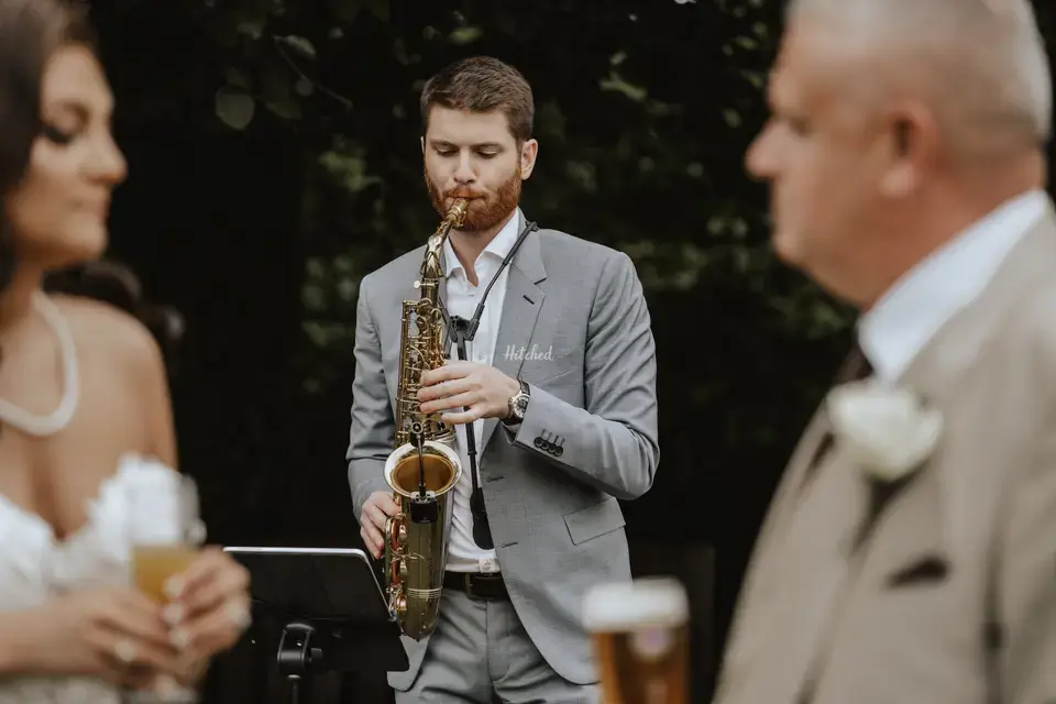 Wedding saxophonist James Burwell playing saxophone in the background at a wedding - the bride and groom are visible but out of focus in the foreground