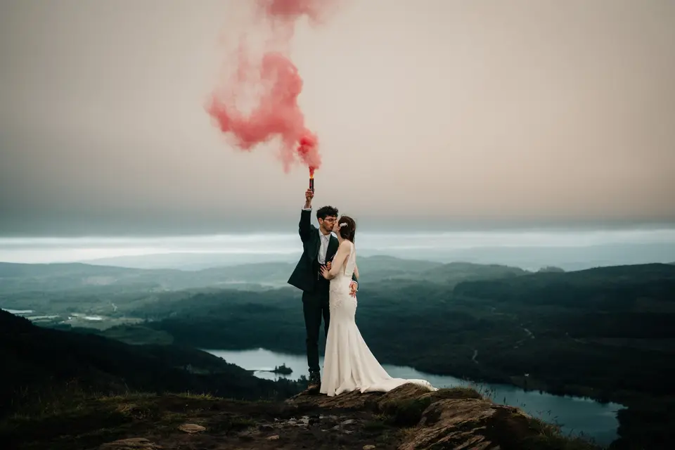 Couple in their wedding attire kissing on top of a hill as the groom holds a pink smoke flare aloft