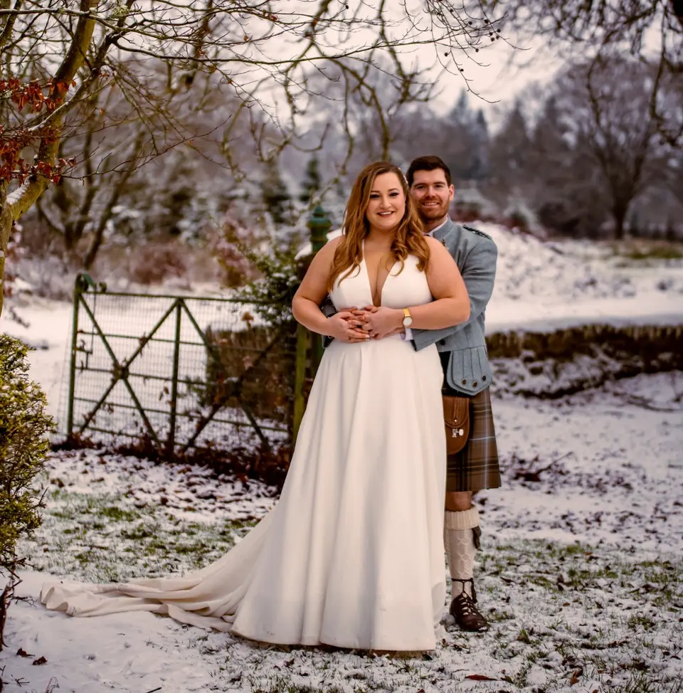 Woman in a wedding dress and a man in a traditional Scottish kilt and formal attire standing together in the snow on their wedding day