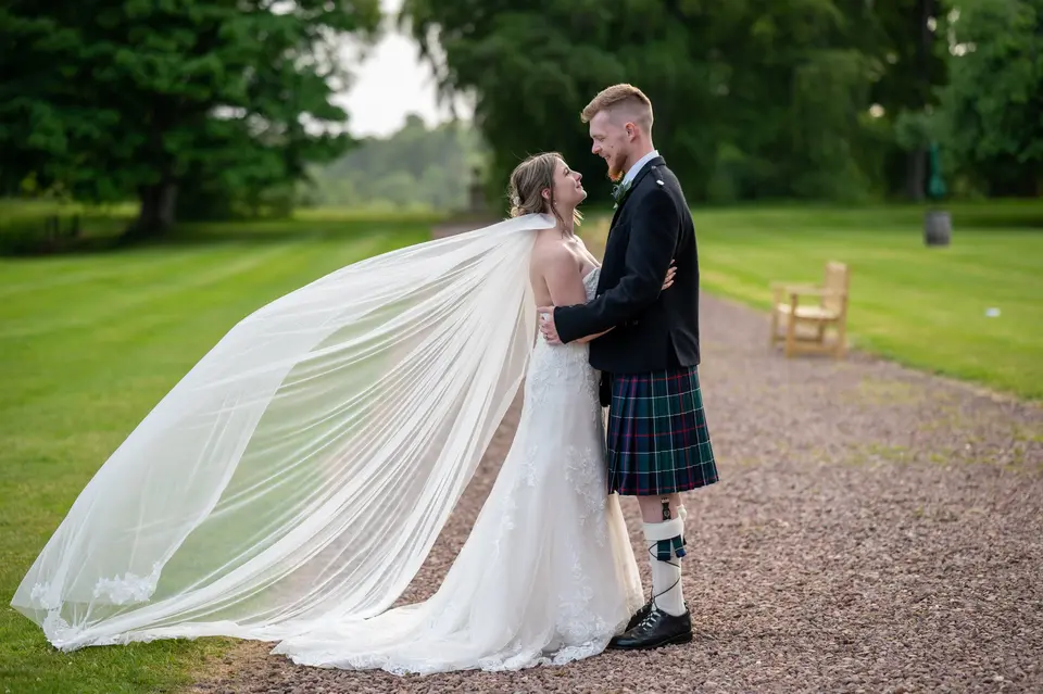 Bride in a wedding dress with her veil blowing in the wind looking up at her groom who is wearing a traditional scottish kilt
