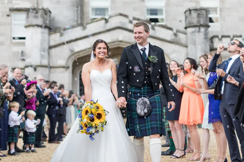Bride and groom in a traditional scottish kilt and formalwear beaming hand in hand as they exit their wedding ceremony, through a crowd of their guests all blowing bubbles