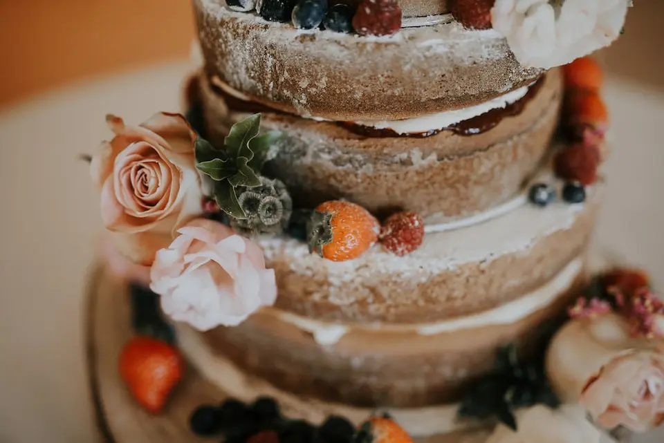 A close-up shot of a rustic naked wedding cake with flowers, real fruit berries and white icing sugar as decoration